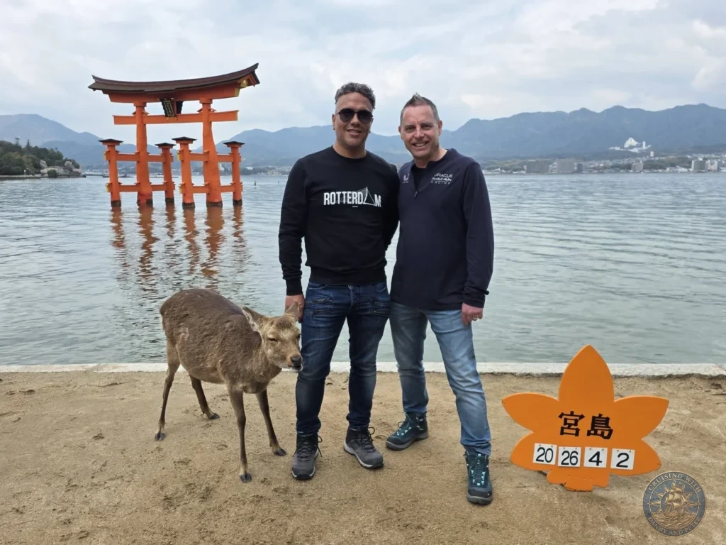 Hiroshima Japan Cruise Port Guide 2026 – Insider Tips & One Day Itinerary 2 Danny and Peter standing in front of the iconic floating vermilion Torii Gate with a friendly wild sika deer and date sign on Miyajima Island in Hiroshima Japan. Hiroshima Japan Cruise Port Guide, DIY Hiroshima Cruise Port to City Center, Things to do in Hiroshima from cruise ship, Westerdam Hiroshima Docking Location, One day in Hiroshima Japan itinerary by Danny and Peter.