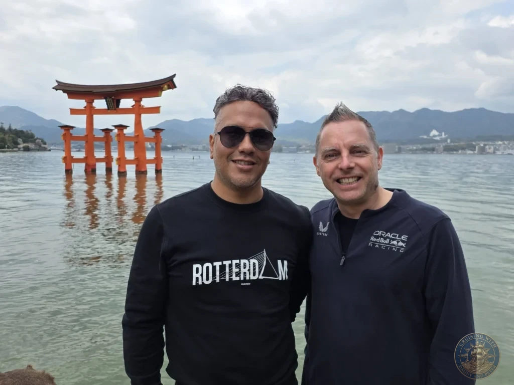 Hiroshima Japan Cruise Port Guide 2026 – Insider Tips & One Day Itinerary 10 Danny and Peter smiling in a close-up picture in front of the floating Grand Torii Gate of Itsukushima Shrine on Miyajima Island in Hiroshima Japan. Hiroshima Japan Cruise Port Guide, DIY Hiroshima Cruise Port to City Center, Things to do in Hiroshima from cruise ship, Westerdam Hiroshima Docking Location, One day in Hiroshima Japan itinerary by Danny and Peter.