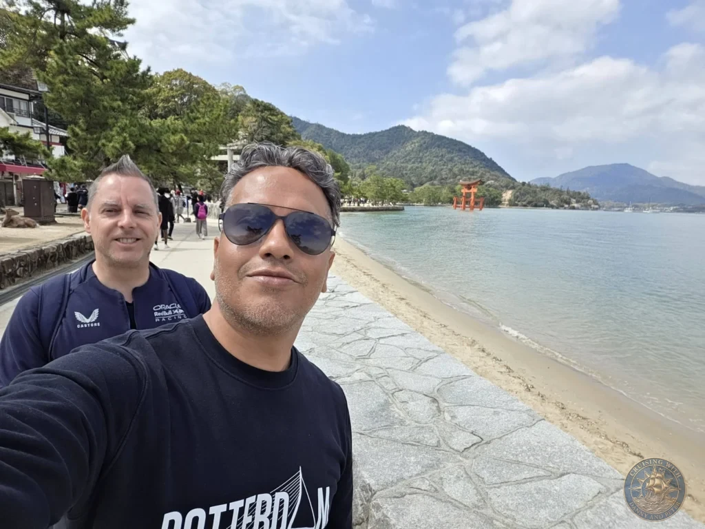 Hiroshima Japan Cruise Port Guide 2026 – Insider Tips & One Day Itinerary 20 Danny and Peter taking a selfie along the coastal stone promenade of Miyajima Island with the iconic floating Torii Gate and sea in the background in Hiroshima Japan. Hiroshima Japan Cruise Port Guide, DIY Hiroshima Cruise Port to City Center, Things to do in Hiroshima from cruise ship, Westerdam Hiroshima Docking Location, One day in Hiroshima Japan itinerary by Danny and Peter.
