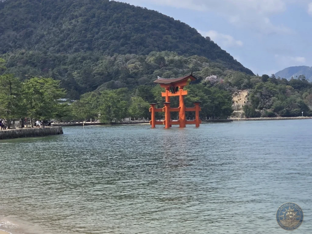 Hiroshima Japan Cruise Port Guide 2026 – Insider Tips & One Day Itinerary 6 Side view of the grand vermilion floating Torii Gate standing in the water near the shore of Miyajima Island framed by lush green hills in Hiroshima Japan. Hiroshima Japan Cruise Port Guide, DIY Hiroshima Cruise Port to City Center, Things to do in Hiroshima from cruise ship, Westerdam Hiroshima Docking Location, One day in Hiroshima Japan itinerary by Danny and Peter.