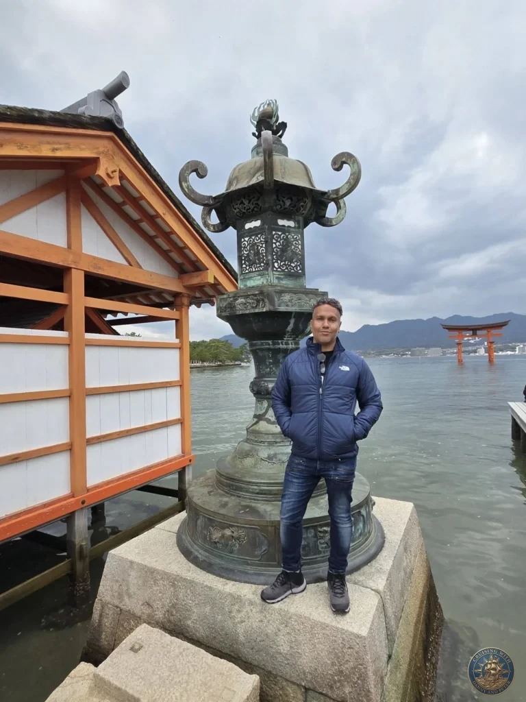 Hiroshima Japan Cruise Port Guide 2026 – Insider Tips & One Day Itinerary 31 Danny posing next to a giant ornate bronze lantern on the wooden boardwalk of Itsukushima Shrine with the floating Torii Gate in the background in Hiroshima Japan. Hiroshima Japan Cruise Port Guide, DIY Hiroshima Cruise Port to City Center, Things to do in Hiroshima from cruise ship, Westerdam Hiroshima Docking Location, One day in Hiroshima Japan itinerary by Danny and Peter.