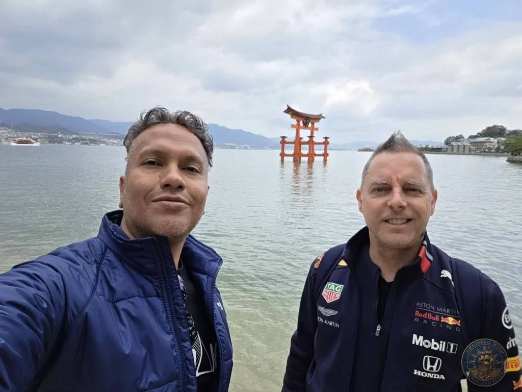Hiroshima Japan Cruise Port Guide 2026 – Insider Tips & One Day Itinerary 73 Danny and Peter smiling for a selfie along the Miyajima shoreline with the floating Grand Torii Gate centered behind them in Hiroshima Japan. Hiroshima Japan Cruise Port Guide, DIY Hiroshima Cruise Port to City Center, Things to do in Hiroshima from cruise ship, Westerdam Hiroshima Docking Location, One day in Hiroshima Japan itinerary by Danny and Peter.