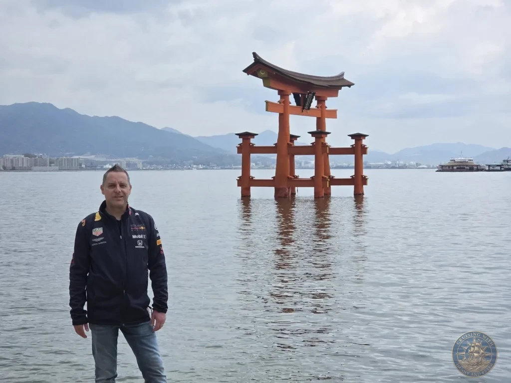Hiroshima Japan Cruise Port Guide 2026 – Insider Tips & One Day Itinerary 32 Peter posing along the Miyajima shoreline with the floating Grand Torii Gate standing in the sea behind him at high tide in Hiroshima Japan. Hiroshima Japan Cruise Port Guide, DIY Hiroshima Cruise Port to City Center, Things to do in Hiroshima from cruise ship, Westerdam Hiroshima Docking Location, One day in Hiroshima Japan itinerary by Danny and Peter.