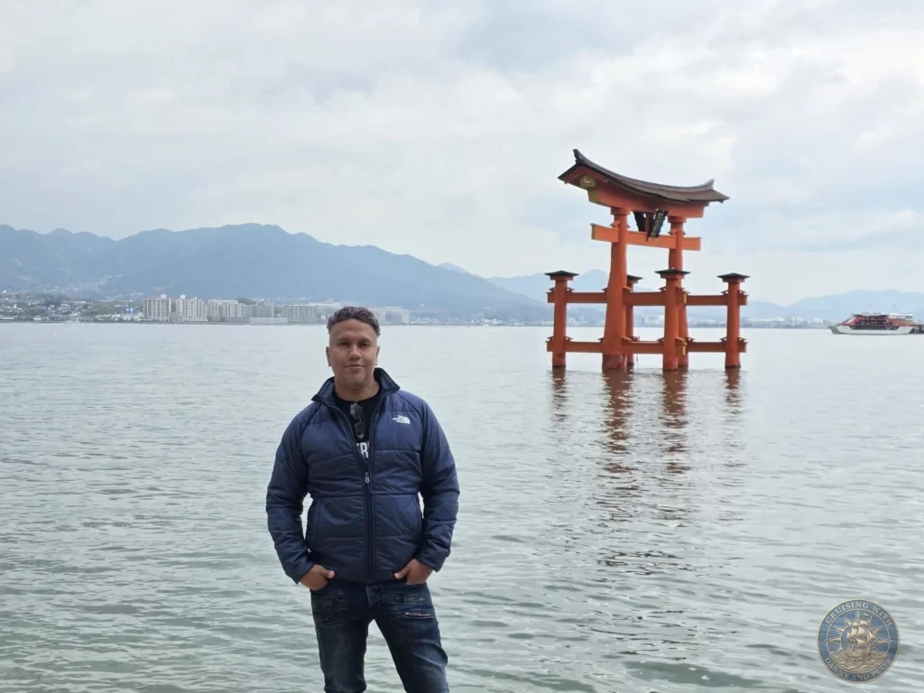 Hiroshima Japan Cruise Port Guide 2026 – Insider Tips & One Day Itinerary 24 Danny standing along the Miyajima waterfront with the Grand Torii Gate rising out of the water at high tide in Hiroshima Japan. Hiroshima Japan Cruise Port Guide, DIY Hiroshima Cruise Port to City Center, Things to do in Hiroshima from cruise ship, Westerdam Hiroshima Docking Location, One day in Hiroshima Japan itinerary by Danny and Peter.
