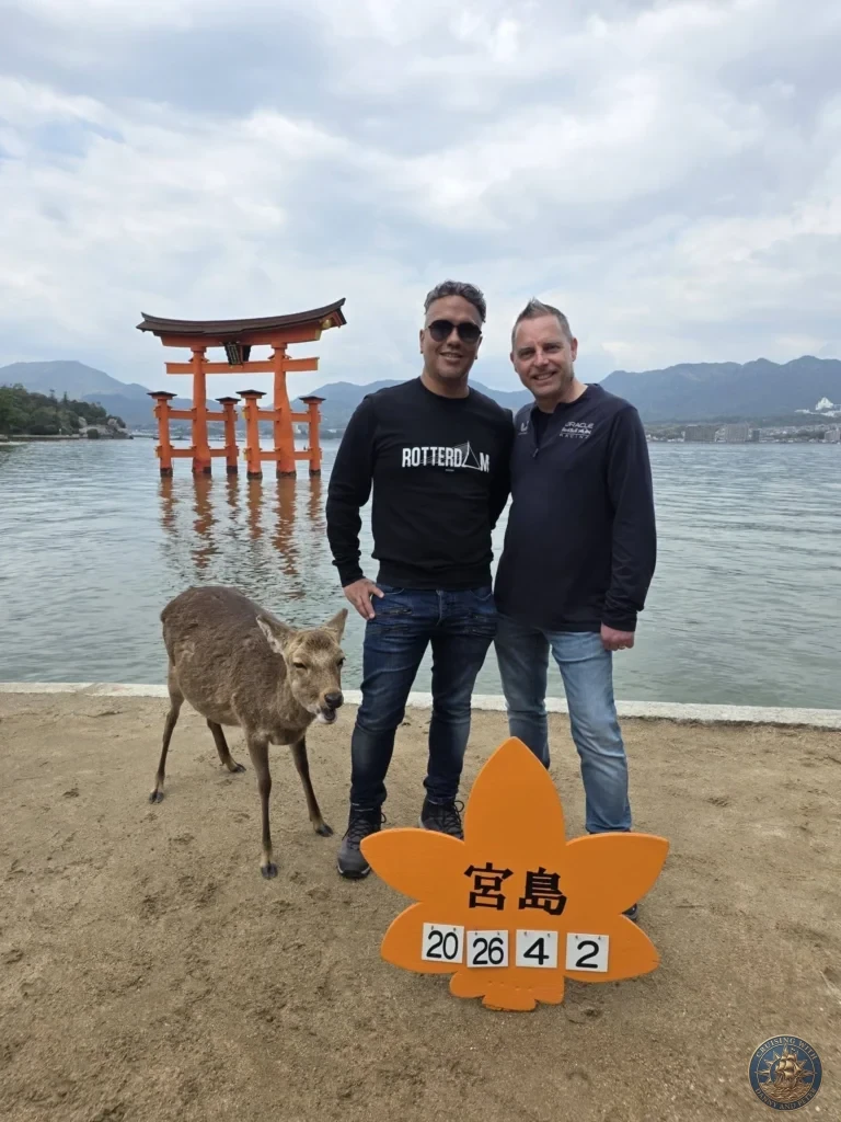 Hiroshima Japan Cruise Port Guide 2026 – Insider Tips & One Day Itinerary 33 Danny and Peter standing next to a wild Sika deer on the Miyajima beach with the Grand Torii Gate and a date sign in the foreground in Hiroshima Japan. Hiroshima Japan Cruise Port Guide, DIY Hiroshima Cruise Port to City Center, Things to do in Hiroshima from cruise ship, Westerdam Hiroshima Docking Location, One day in Hiroshima Japan itinerary by Danny and Peter.