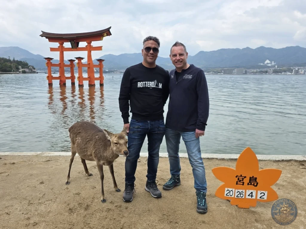 Hiroshima Japan Cruise Port Guide 2026 – Insider Tips & One Day Itinerary 14 Danny and Peter standing on the sandy shoreline of Miyajima with a wild Sika deer, the Grand Torii Gate floating at high tide, and a orange maple leaf date sign in Hiroshima Japan. Hiroshima Japan Cruise Port Guide, DIY Hiroshima Cruise Port to City Center, Things to do in Hiroshima from cruise ship, Westerdam Hiroshima Docking Location, One day in Hiroshima Japan itinerary by Danny and Peter.