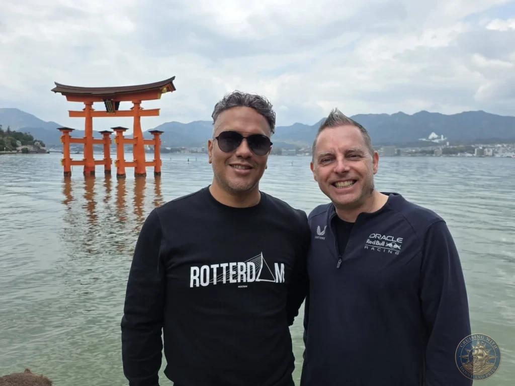 Hiroshima Japan Cruise Port Guide 2026 – Insider Tips & One Day Itinerary 78 Danny and Peter smiling for a portrait along the Miyajima shoreline with the floating Grand Torii Gate standing in the sea behind them in Hiroshima Japan. Hiroshima Japan Cruise Port Guide, DIY Hiroshima Cruise Port to City Center, Things to do in Hiroshima from cruise ship, Westerdam Hiroshima Docking Location, One day in Hiroshima Japan itinerary by Danny and Peter.