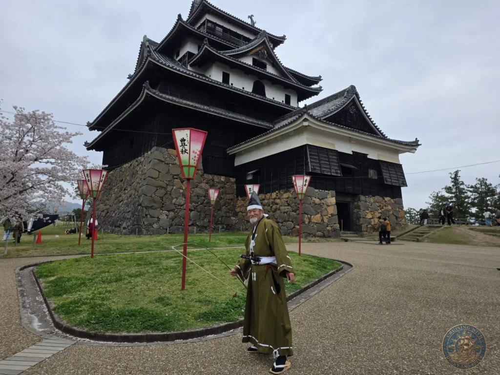 A local Japanese performer dressed in traditional olive-green attire and cap standing in front of the historic Matsue Castle keep decorated with paper lanterns during a shore excursion from Sakaiminato Japan Cruise Port Guide, DIY Sakaiminato Cruise Port to City Center, Things to do in Sakaiminato from cruise ship, Westerdam Sakaiminato Docking Location, One day in Sakaiminato Japan itinerary by Danny and Peter.
