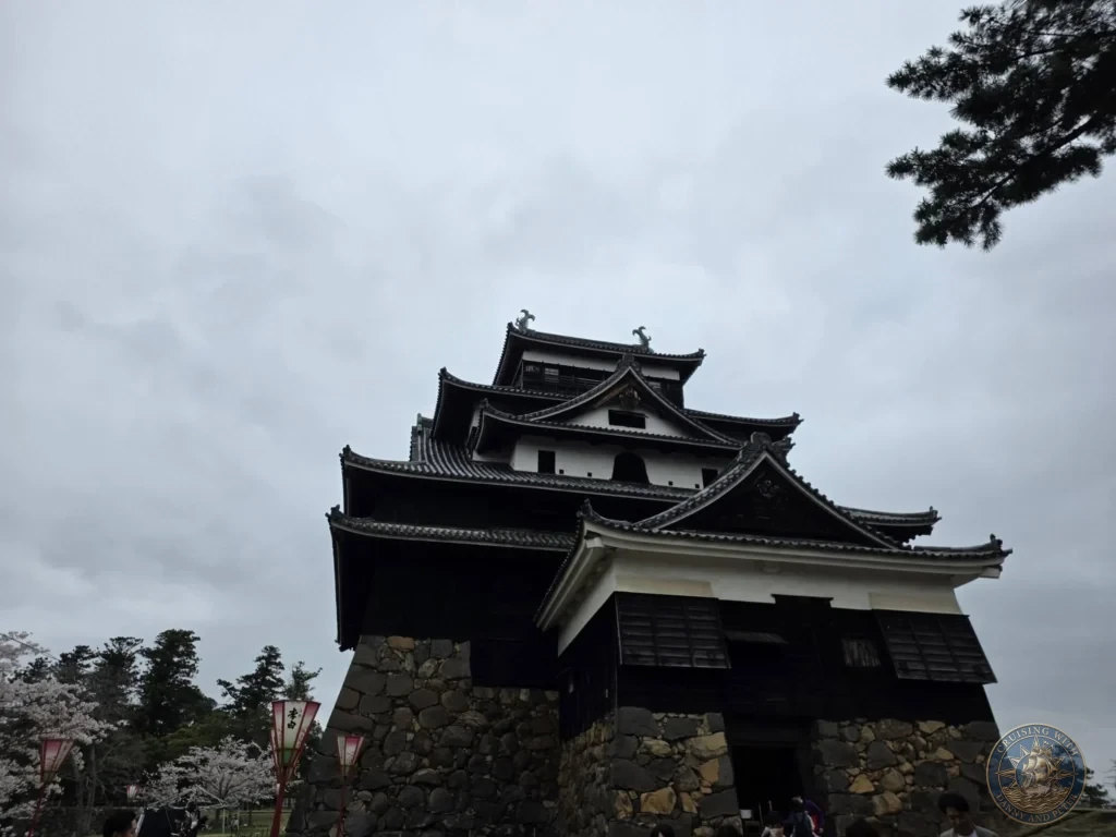 A low-angle view looking up at the historic black wooden main keep of Matsue Castle with stone foundations and paper lanterns during a shore excursion from Sakaiminato Japan Cruise Port Guide, DIY Sakaiminato Cruise Port to City Center, Things to do in Sakaiminato from cruise ship, Westerdam Sakaiminato Docking Location, One day in Sakaiminato Japan itinerary by Danny and Peter.