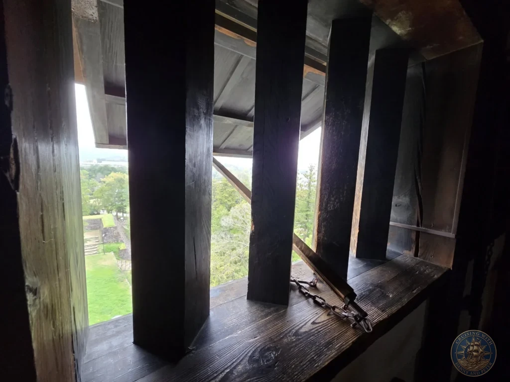 Looking out through heavy wooden defensive window grates and thick wooden frames inside Matsue Castle keep toward the green park grounds below during a shore excursion from Sakaiminato Japan Cruise Port Guide, DIY Sakaiminato Cruise Port to City Center, Things to do in Sakaiminato from cruise ship, Westerdam Sakaiminato Docking Location, One day in Sakaiminato Japan itinerary by Danny and Peter.