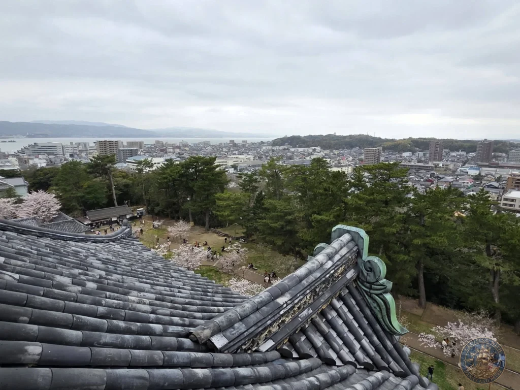 High-angle perspective from the top of Matsue Castle looking over intricate grey roof tiles and green gable ornaments toward Lake Shinji, the city skyline, and cherry blossom trees during a shore excursion from Sakaiminato Japan Cruise Port Guide, DIY Sakaiminato Cruise Port to City Center, Things to do in Sakaiminato from cruise ship, Westerdam Sakaiminato Docking Location, One day in Sakaiminato Japan itinerary by Danny and Peter.