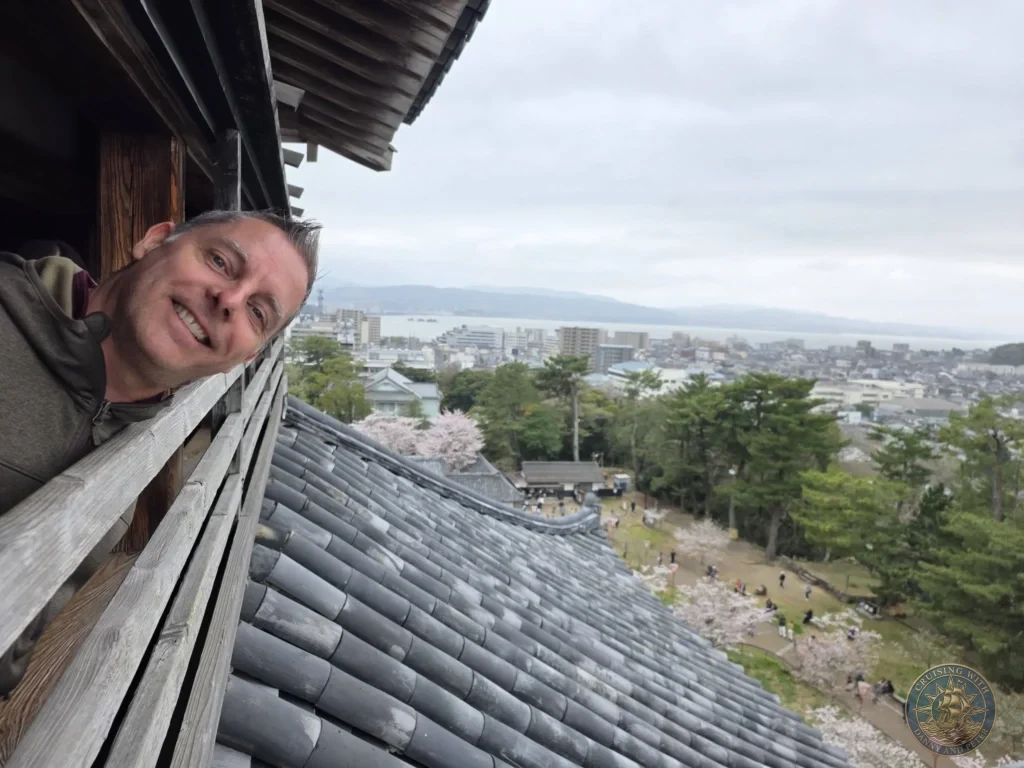 Peter smiling while leaning out from an open wooden window on the top floor of Matsue Castle keep with tiled roofs, cherry blossoms, and Lake Shinji in the background during a shore excursion from Sakaiminato Japan Cruise Port Guide, DIY Sakaiminato Cruise Port to City Center, Things to do in Sakaiminato from cruise ship, Westerdam Sakaiminato Docking Location, One day in Sakaiminato Japan itinerary by Danny and Peter.
