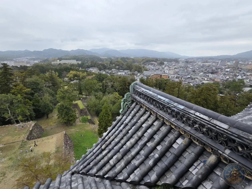 High-angle view looking down over the intricate tiled roof ridge and stone foundation walls of Matsue Castle toward forested hills and distant mountains during a shore excursion from Sakaiminato Japan Cruise Port Guide, DIY Sakaiminato Cruise Port to City Center, Things to do in Sakaiminato from cruise ship, Westerdam Sakaiminato Docking Location, One day in Sakaiminato Japan itinerary by Danny and Peter.