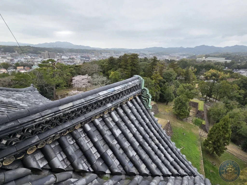 Close-up perspective of the ornate curved roof tile ridge and ornamental green gable end of Matsue Castle looking out over surrounding greenery and distant mountains during a shore excursion from Sakaiminato Japan Cruise Port Guide, DIY Sakaiminato Cruise Port to City Center, Things to do in Sakaiminato from cruise ship, Westerdam Sakaiminato Docking Location, One day in Sakaiminato Japan itinerary by Danny and Peter.