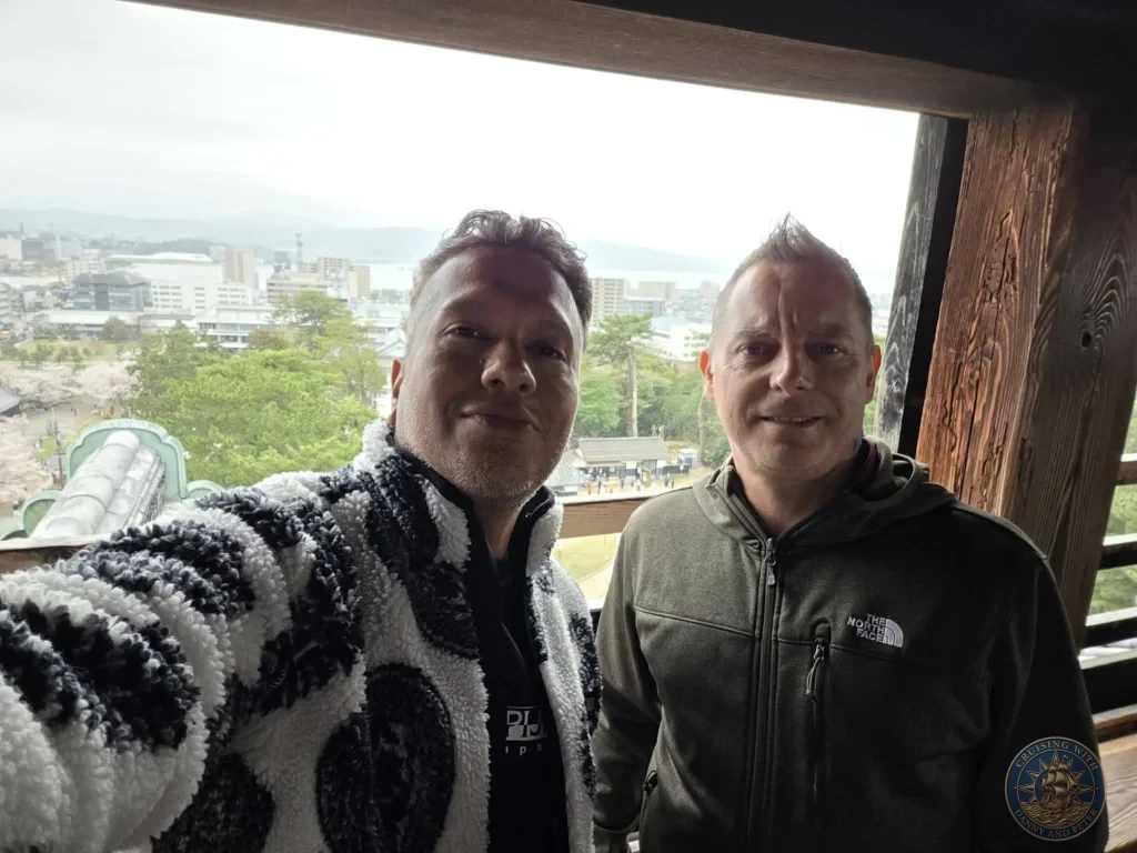 Danny and Peter smiling together inside the top floor of Matsue Castle keep with Lake Shinji, cherry blossoms, and the city of Matsue visible through the wooden window frame during a shore excursion from Sakaiminato Japan Cruise Port Guide, DIY Sakaiminato Cruise Port to City Center, Things to do in Sakaiminato from cruise ship, Westerdam Sakaiminato Docking Location, One day in Sakaiminato Japan itinerary by Danny and Peter.