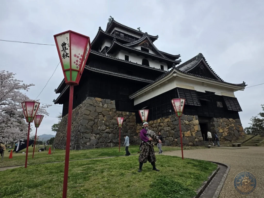 Wide view of the historic black wood Matsue Castle keep and stone wall foundation with a local performer in traditional samurai costume standing on the lawn surrounded by festival paper lanterns during a shore excursion from Sakaiminato Japan Cruise Port Guide, DIY Sakaiminato Cruise Port to City Center, Things to do in Sakaiminato from cruise ship, Westerdam Sakaiminato Docking Location, One day in Sakaiminato Japan itinerary by Danny and Peter.
