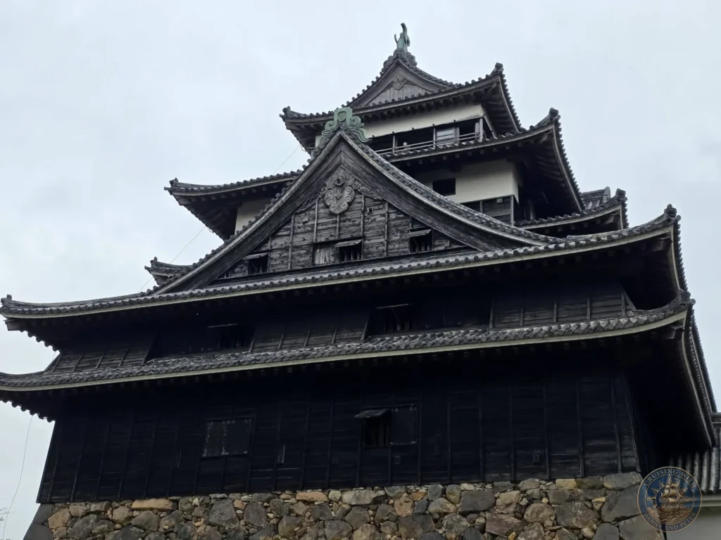 Low angle view of the multi-tiered dark wood exterior and stone foundation of Matsue Castle main keep against an overcast sky during a shore excursion from Sakaiminato Japan Cruise Port Guide, DIY Sakaiminato Cruise Port to City Center, Things to do in Sakaiminato from cruise ship, Westerdam Sakaiminato Docking Location, One day in Sakaiminato Japan itinerary by Danny and Peter.