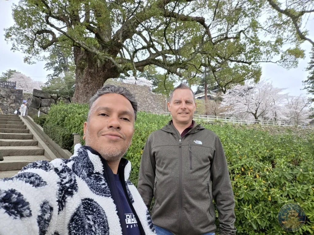 Selfie of Danny and Peter smiling in front of the stone staircases, lush green trees, and blooming cherry blossoms on the grounds of Matsue Castle during a shore excursion from Sakaiminato Japan Cruise Port Guide, DIY Sakaiminato Cruise Port to City Center, Things to do in Sakaiminato from cruise ship, Westerdam Sakaiminato Docking Location, One day in Sakaiminato Japan itinerary by Danny and Peter.