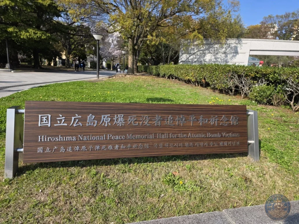 Hiroshima Japan Cruise Port Guide 2026 – Insider Tips & One Day Itinerary 65 A wooden entrance sign for the Hiroshima National Peace Memorial Hall for the Atomic Bomb Victims surrounded by green grass and trees in Hiroshima Japan. Hiroshima Japan Cruise Port Guide, DIY Hiroshima Cruise Port to City Center, Things to do in Hiroshima from cruise ship, Westerdam Hiroshima Docking Location, One day in Hiroshima Japan itinerary by Danny and Peter.