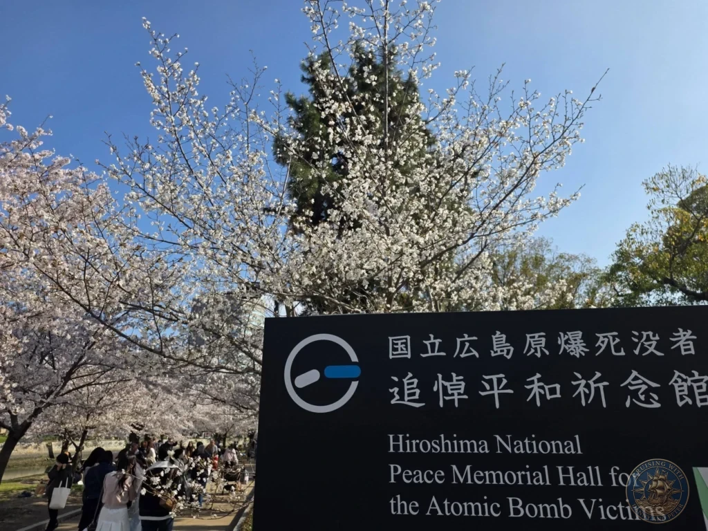 Hiroshima Japan Cruise Port Guide 2026 – Insider Tips & One Day Itinerary 67 Cherry blossom trees in full bloom framing the sign for the Hiroshima National Peace Memorial Hall for the Atomic Bomb Victims under a bright blue sky in Hiroshima Japan. Hiroshima Japan Cruise Port Guide, DIY Hiroshima Cruise Port to City Center, Things to do in Hiroshima from cruise ship, Westerdam Hiroshima Docking Location, One day in Hiroshima Japan itinerary by Danny and Peter.