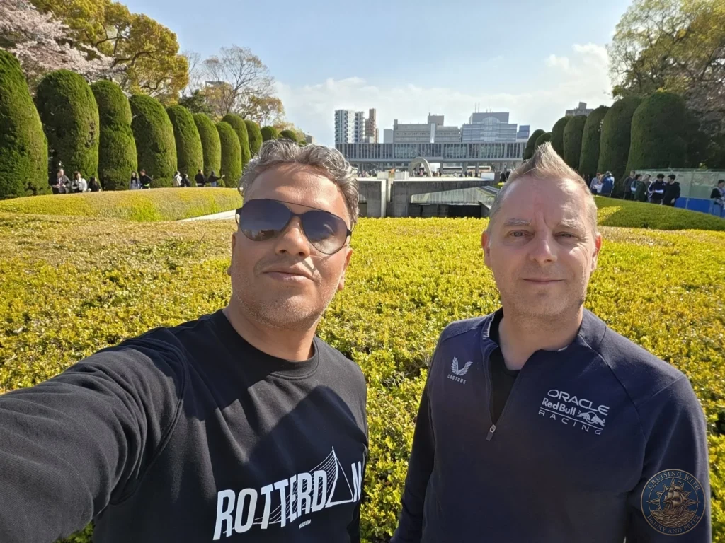 Hiroshima Japan Cruise Port Guide 2026 – Insider Tips & One Day Itinerary 5 A selfie of Danny and Peter standing in front of manicured hedges with the Hiroshima Peace Memorial Museum in the background under a sunny blue sky in Hiroshima Japan. Hiroshima Japan Cruise Port Guide, DIY Hiroshima Cruise Port to City Center, Things to do in Hiroshima from cruise ship, Westerdam Hiroshima Docking Location, One day in Hiroshima Japan itinerary by Danny and Peter.