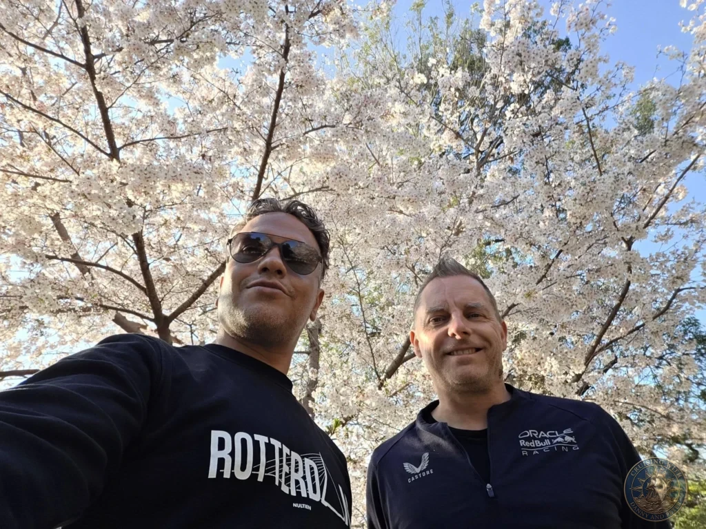 Hiroshima Japan Cruise Port Guide 2026 – Insider Tips & One Day Itinerary 13 A low-angle selfie of Danny and Peter standing beneath a full canopy of blooming pink cherry blossoms under a clear blue sky in Peace Memorial Park in Hiroshima Japan. Hiroshima Japan Cruise Port Guide, DIY Hiroshima Cruise Port to City Center, Things to do in Hiroshima from cruise ship, Westerdam Hiroshima Docking Location, One day in Hiroshima Japan itinerary by Danny and Peter.
