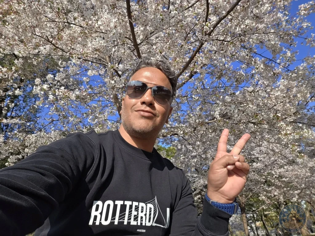Hiroshima Japan Cruise Port Guide 2026 – Insider Tips & One Day Itinerary 23 A close-up selfie displaying a peace sign gesture framed by a vibrant, dense canopy of pale pink cherry blossoms blooming under a clear blue sky in Peace Memorial Park in Hiroshima Japan. Hiroshima Japan Cruise Port Guide, DIY Hiroshima Cruise Port to City Center, Things to do in Hiroshima from cruise ship, Westerdam Hiroshima Docking Location, One day in Hiroshima Japan itinerary by Danny and Peter.