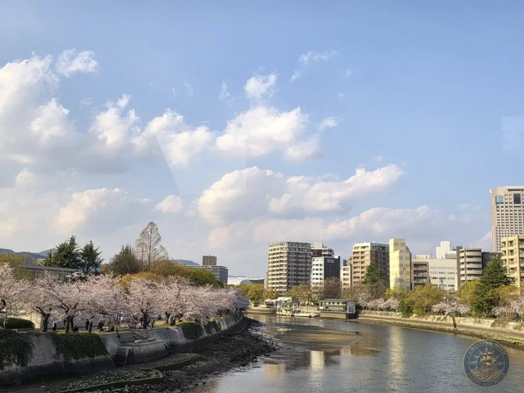 Hiroshima Japan Cruise Port Guide 2026 – Insider Tips & One Day Itinerary 9 A scenic wide landscape view showing the Motoyasu River lined with blooming pink cherry blossom trees leading toward modern city buildings under a partly cloudy sky at Peace Memorial Park in Hiroshima Japan. Hiroshima Japan Cruise Port Guide, DIY Hiroshima Cruise Port to City Center, Things to do in Hiroshima from cruise ship, Westerdam Hiroshima Docking Location, One day in Hiroshima Japan itinerary by Danny and Peter.