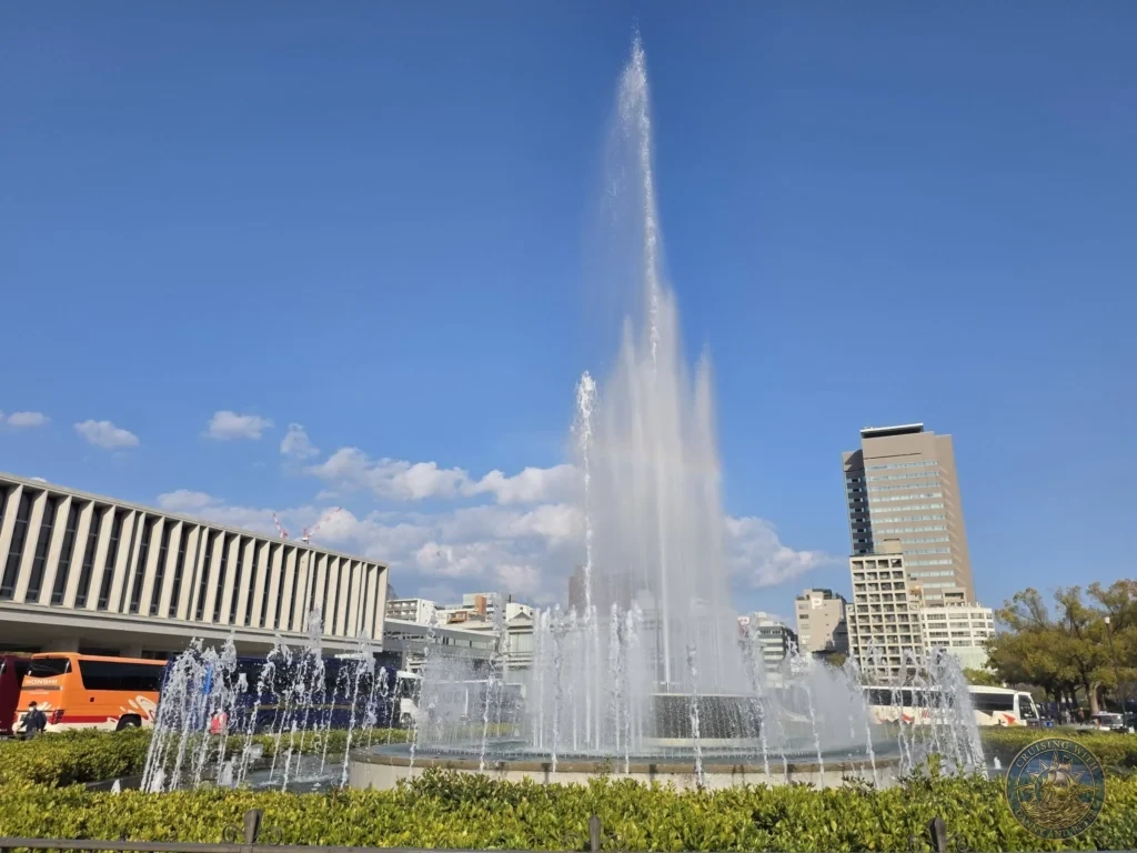 Hiroshima Japan Cruise Port Guide 2026 – Insider Tips & One Day Itinerary 25 A vibrant water fountain shooting high jets into the clear blue sky in front of the Hiroshima Peace Memorial Museum in Hiroshima Japan. Hiroshima Japan Cruise Port Guide, DIY Hiroshima Cruise Port to City Center, Things to do in Hiroshima from cruise ship, Westerdam Hiroshima Docking Location, One day in Hiroshima Japan itinerary by Danny and Peter.