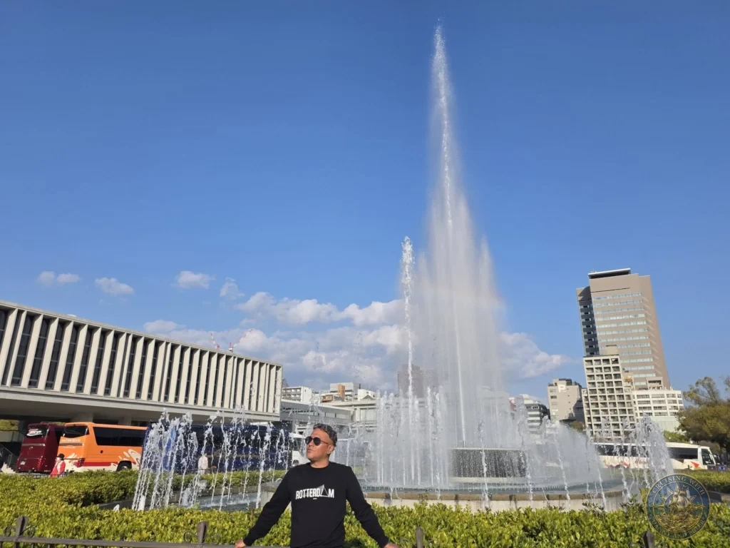 Hiroshima Japan Cruise Port Guide 2026 – Insider Tips & One Day Itinerary 75 Danny standing in front of the Fountain of Prayer as high water jets spray under a blue sky near the Peace Memorial Museum in Hiroshima Japan. Hiroshima Japan Cruise Port Guide, DIY Hiroshima Cruise Port to City Center, Things to do in Hiroshima from cruise ship, Westerdam Hiroshima Docking Location, One day in Hiroshima Japan itinerary by Danny and Peter.