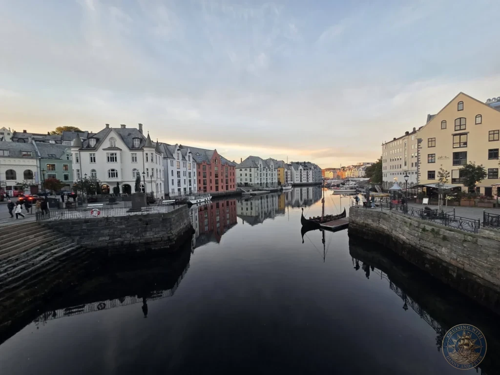 Brosundet canal lined with historic Art Nouveau architecture, calm waters reflecting colorful waterfront buildings, and a docked traditional boat in Ålesund, Norway. DIY Ålesund Cruise Port to City Center, Things to do in Ålesund from cruise ship, Rotterdam Ålesund Docking Location, One day in Ålesund Norway itinerary, by Danny and Peter.