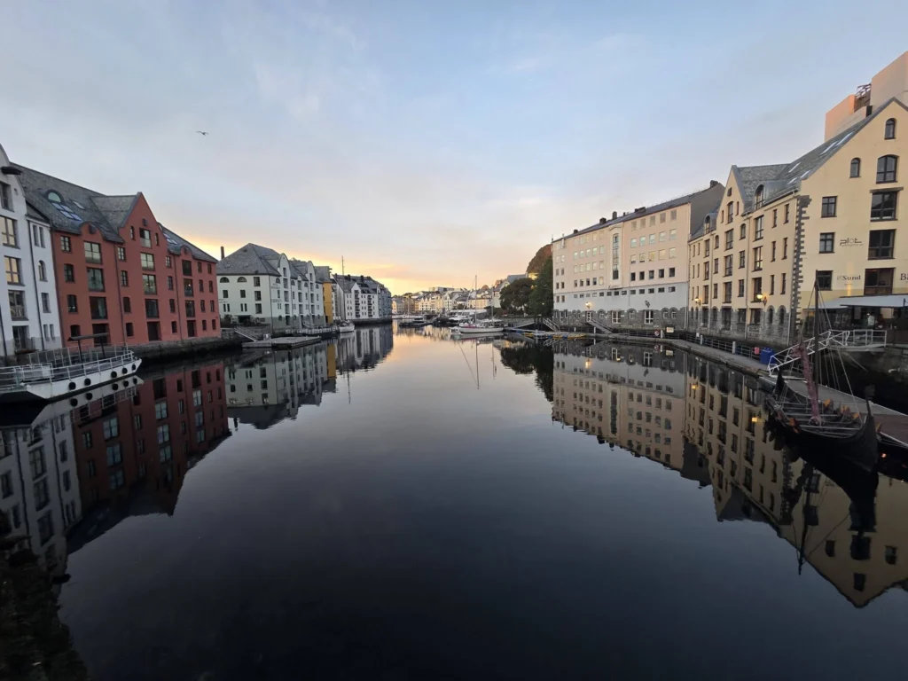 A wide-angle landscape shot captures the smooth waters of the Brosundet canal at dawn or dusk in Ålesund, perfectly reflecting the pastel and brick Art Nouveau waterfront buildings. The sky is clear, pale blue with hints of peach. A single large bird flies high. A small traditional boat sits by the right bank, and other small vessels are further down the waterway. This image is part of our Ålesund Norway Cruise Port Guide and highlights DIY Ålesund Cruise Port to City Center access. Find things to do in Ålesund from cruise ship in our one day itinerary. Also details Rotterdam Ålesund Docking Location, by Danny and Peter.