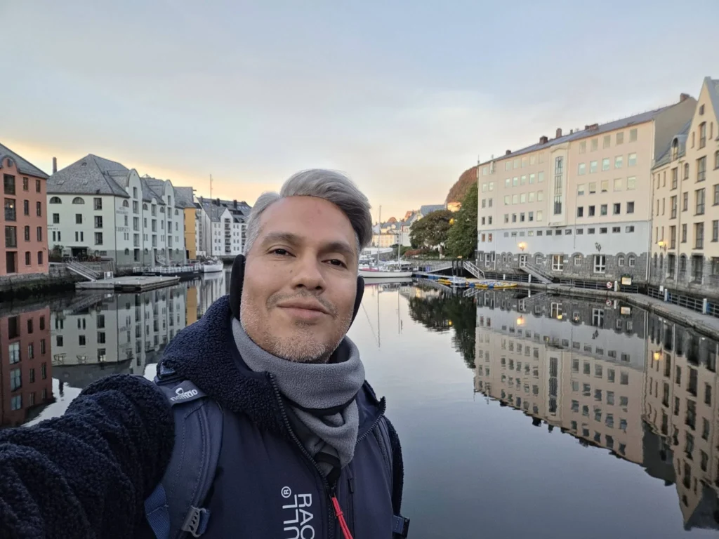 Danny taking a selfie in front of the scenic Brosundet Canal lined with colourful Art Nouveau buildings during a Holland America Rotterdam shore excursion stop. Things to do in Ålesund from cruise ship, by Danny and Peter.