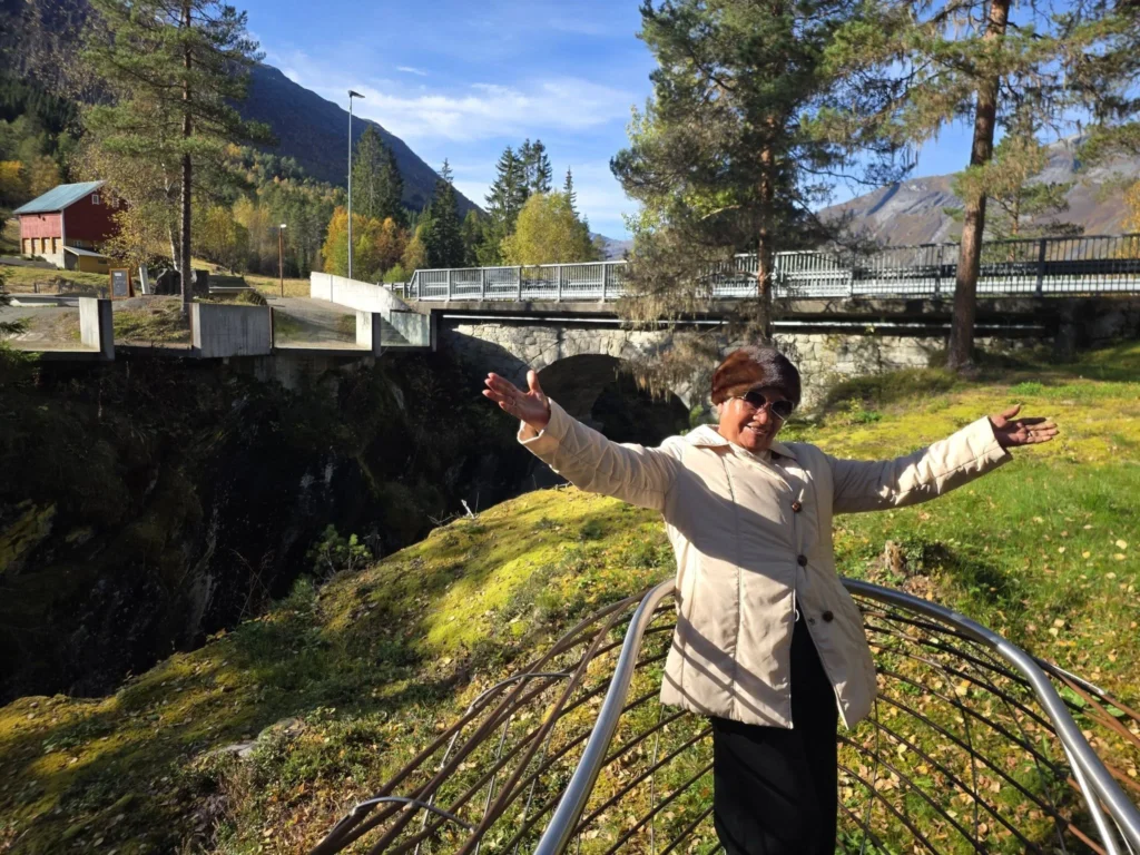 Danny's mother posing joyful with open arms on the modern viewing platform at Gudbrandsjuvet Gorge with bridges and autumn mountains in the background during a Holland America Rotterdam excursion stop. Things to do in Ålesund from cruise ship, by Danny and Peter.