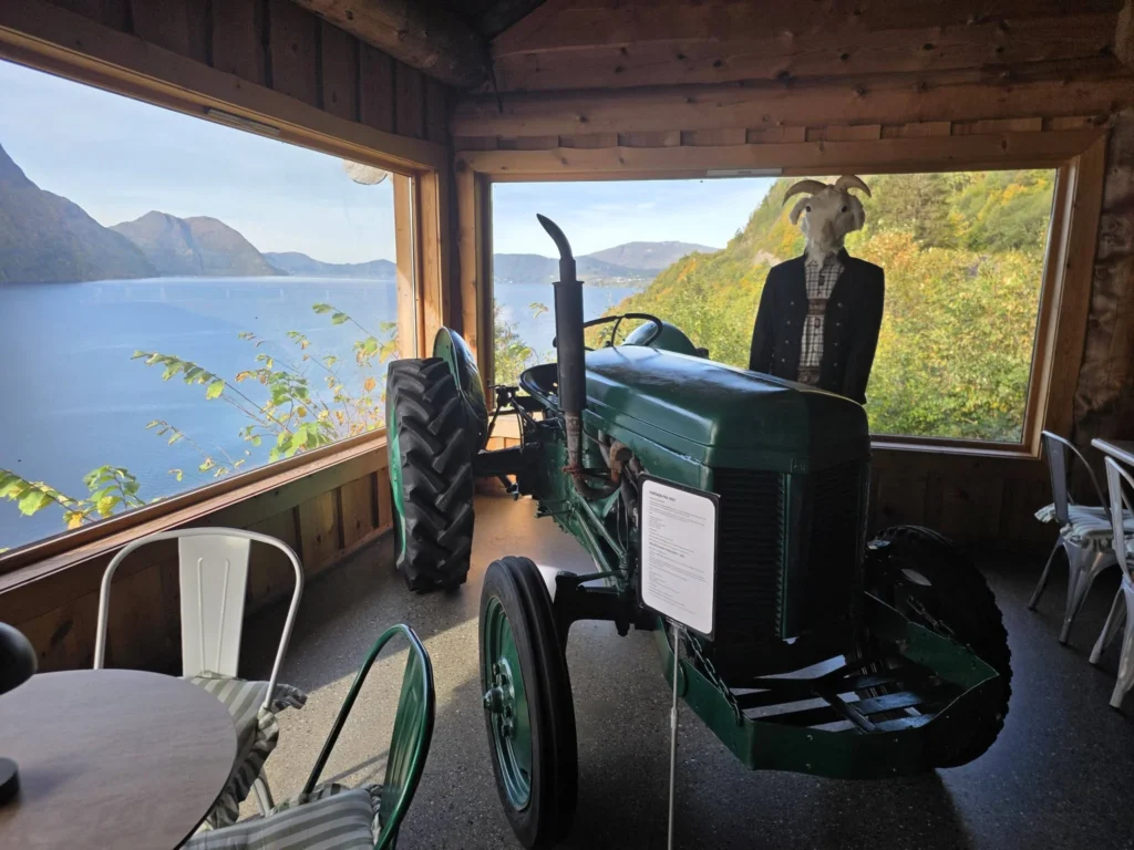 A green vintage tractor and mannequin inside a wooden rest stop with large panoramic windows overlooking the fjord at Kokarsteinen Viewpoint during a Holland America Rotterdam shore excursion. Things to do in Ålesund from cruise ship, by Danny and Peter.