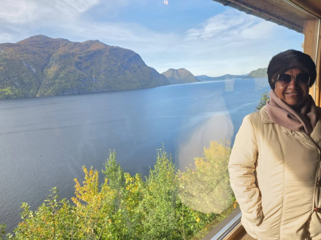 Danny's mother posing beside a large window overlooking the scenic fjord and autumn mountains at Kokarsteinen Viewpoint during a Holland America Rotterdam shore excursion. Things to do in Ålesund from cruise ship, by Danny and Peter.