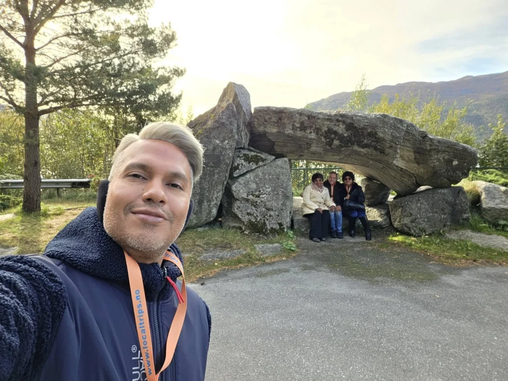 Danny taking a selfie with Peter and family sitting under the large stone arch monument at Kokarsteinen Viewpoint during a Holland America Rotterdam shore excursion. Things to do in Ålesund from cruise ship, by Danny and Peter.