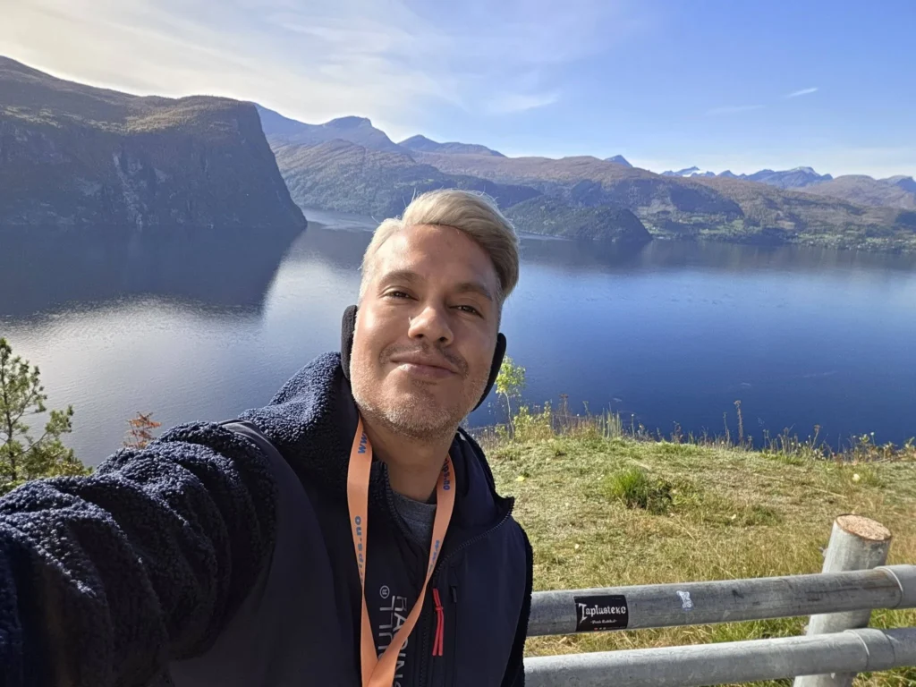 Danny taking a solo selfie overlooking the bright blue fjord and mountain landscape at Kokarsteinen Viewpoint during a Holland America Rotterdam shore excursion. Things to do in Ålesund from cruise ship, by Danny and Peter.