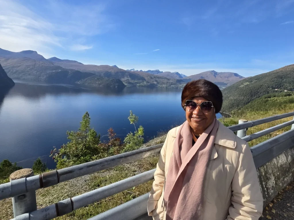 Danny's mother smiling outdoors by the guardrail at Kokarsteinen Viewpoint with deep blue fjord waters and surrounding mountain range behind her during a Holland America Rotterdam shore excursion. Things to do in Ålesund from cruise ship, by Danny and Peter.