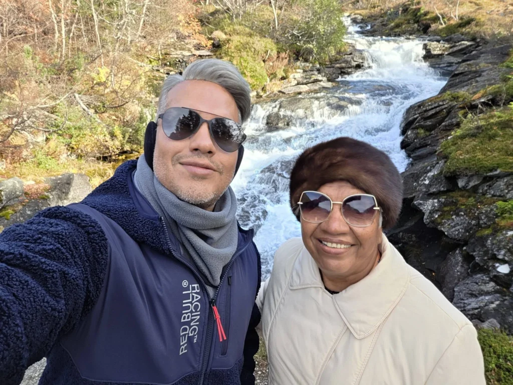 Danny taking a selfie with his mother in front of the cascading mountain stream and waterfall at Krikebrua Waterfall Bridge during a Holland America Rotterdam shore excursion. Things to do in Ålesund from cruise ship, by Danny and Peter.
