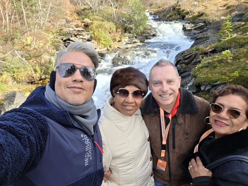 Danny taking a group selfie with his mother, Peter, and family in front of the rushing cascade at Krikebrua Waterfall Bridge during a Holland America Rotterdam shore excursion. Things to do in Ålesund from cruise ship, by Danny and Peter.