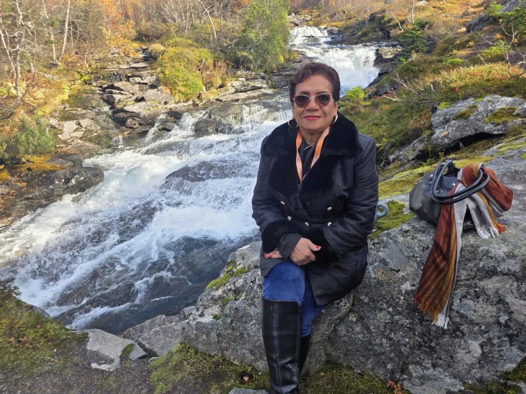 Danny's aunt sitting on a rock beside the rushing waters and cascades of Krikebrua Waterfall Bridge during a Holland America Rotterdam shore excursion. Things to do in Ålesund from cruise ship, by Danny and Peter.