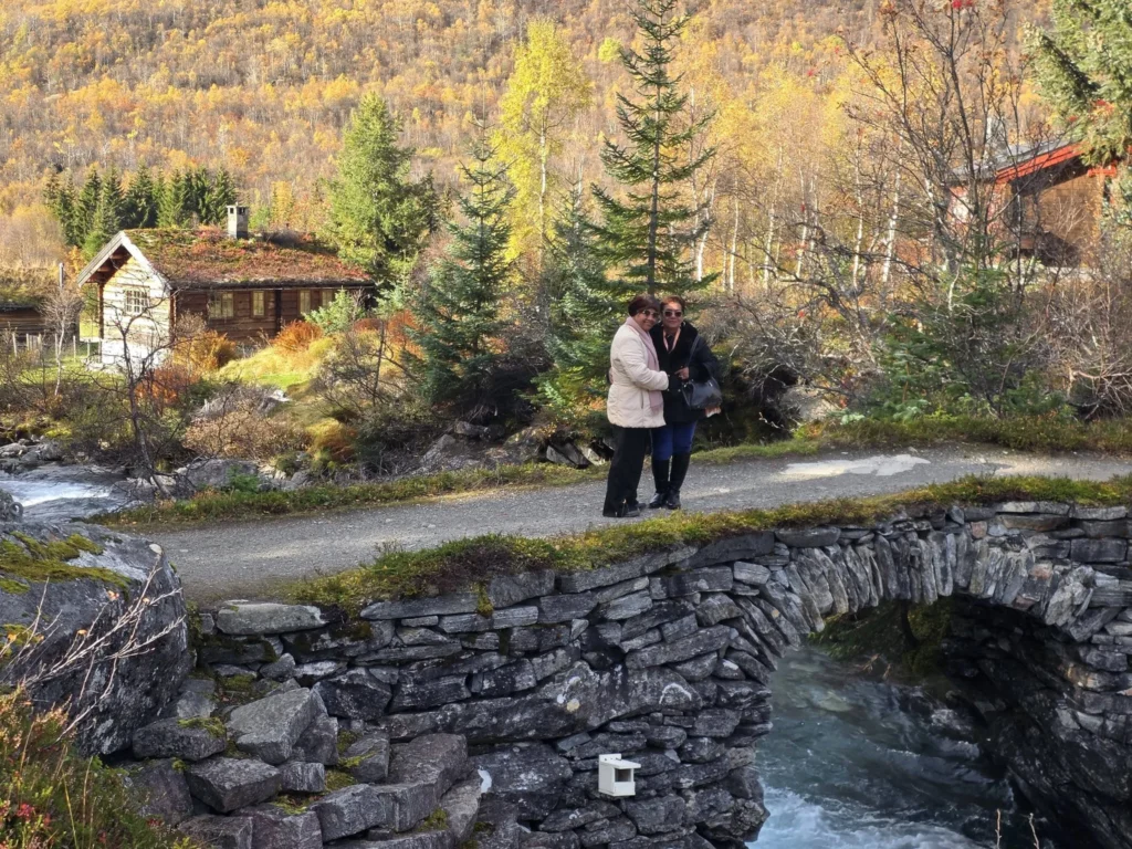 Danny's mother and aunt standing on the historic stone arch bridge at Krikebrua Waterfall, surrounded by turf-roofed wooden cabins and golden autumn mountain scenery during a Holland America Rotterdam shore excursion. Things to do in Ålesund from cruise ship, by Danny and Peter.