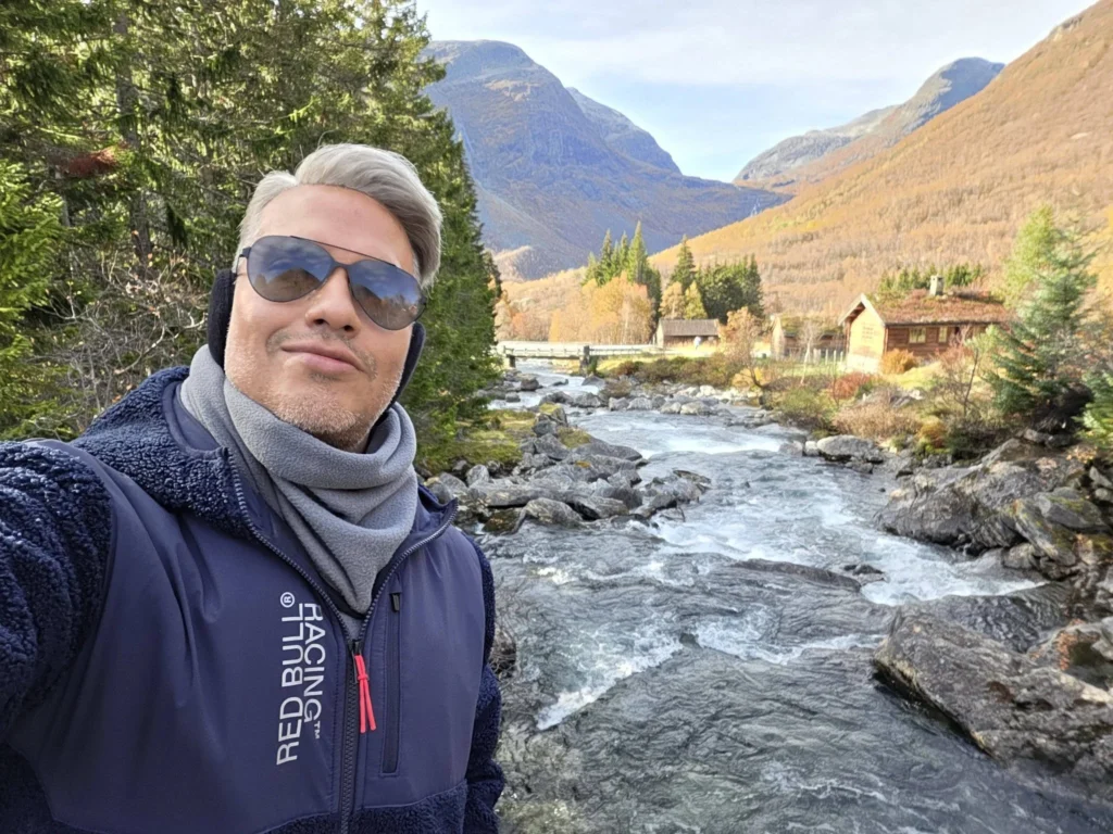 Danny taking a solo selfie along the flowing stream with a stone bridge, grass-roofed wooden cabins, and mountain peaks in the background at Krikebrua Waterfall during a Holland America Rotterdam shore excursion. Things to do in Ålesund from cruise ship, by Danny and Peter.
