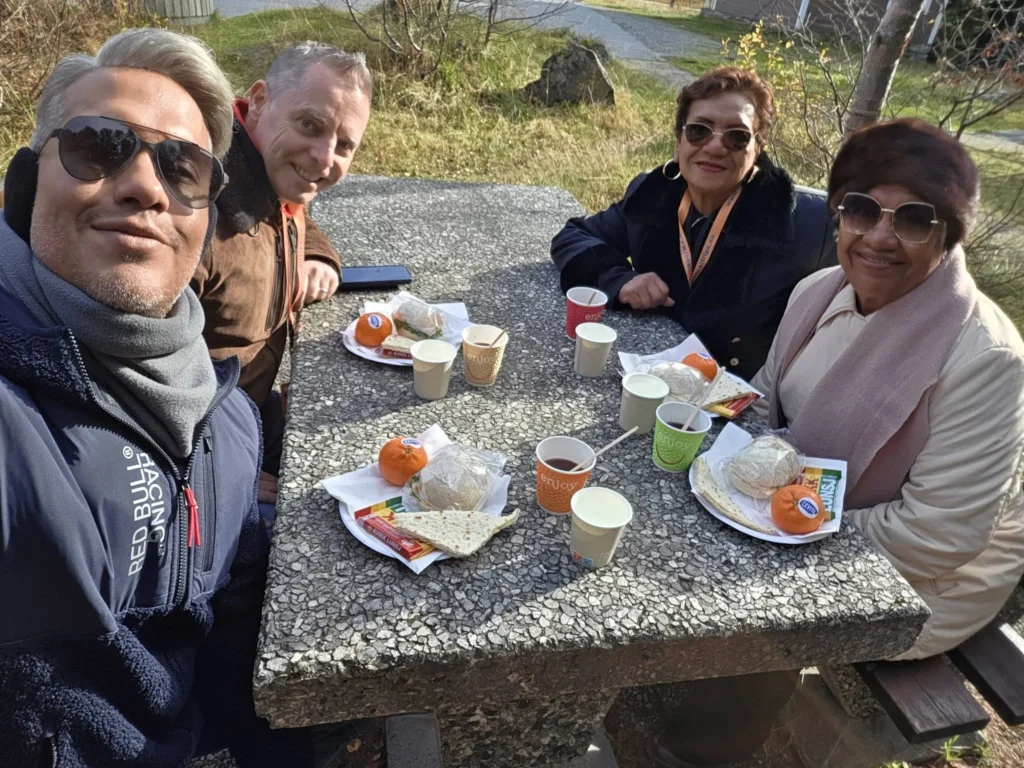 Danny taking a group selfie sitting around a stone picnic table with Peter, his mother, and his aunt, enjoying an outdoor lunch with warm hot cherry juice near Krikebrua Waterfall during a Holland America Rotterdam shore excursion. Things to do in Ålesund from cruise ship, by Danny and Peter.