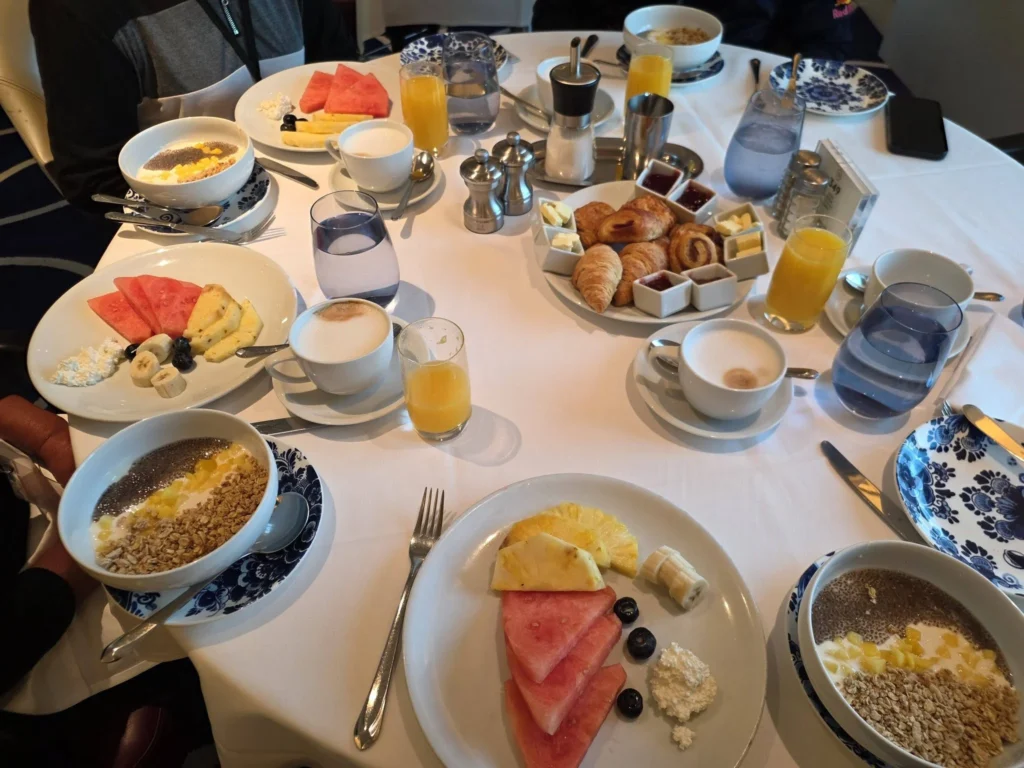 A round dining table spread with fresh watermelon, pineapple, yogurt granola bowls, warm croissants, orange juice, and coffee inside the main dining room onboard Holland America Line's ship Rotterdam during a port call in Ålesund. Things to do in Ålesund from cruise ship, by Danny and Peter.