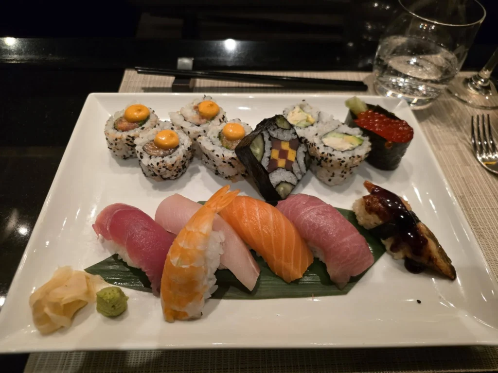 An artfully arranged sushi platter with assorted nigiri, specialty maki rolls, and fresh salmon roe served on a white rectangular plate inside Morimoto by Sea onboard Holland America Line's ship Rotterdam during a cruise to Ålesund. Things to do in Ålesund from cruise ship, by Danny and Peter.