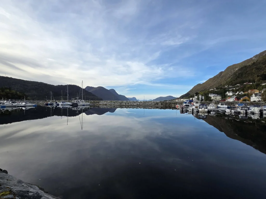 Placid harbor waters perfectly reflecting blue skies, surrounding mountains, and small docked boats along the coastline near Ålesund during a Holland America Rotterdam shore excursion. Things to do in Ålesund from cruise ship, by Danny and Peter.