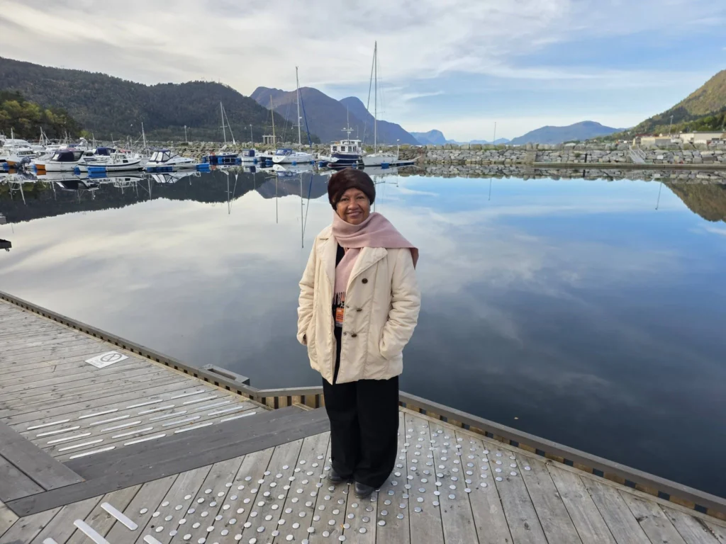Danny's mother standing on a wooden pier in front of a calm harbor with reflections of mountains and boats near Ålesund during a Holland America Rotterdam shore excursion. Things to do in Ålesund from cruise ship, by Danny and Peter.