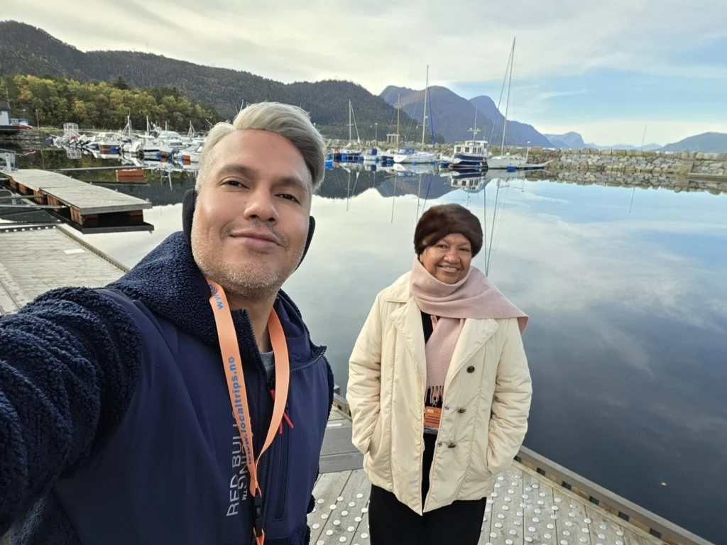 Danny taking a selfie with his mother standing on a wooden pier by a peaceful harbor with sailboat reflections and surrounding mountains near Ålesund during a Holland America Rotterdam shore excursion. Things to do in Ålesund from cruise ship, by Danny and Peter.